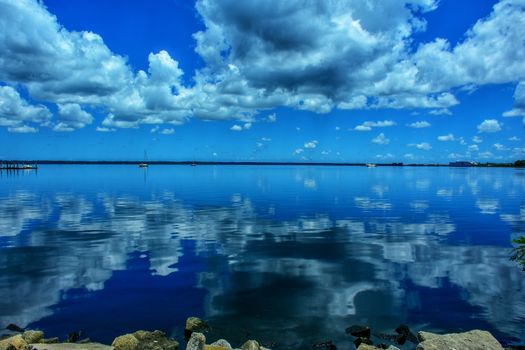 Watery blue expanse, with clouds being reflected in the water and a sailboat in the distance.