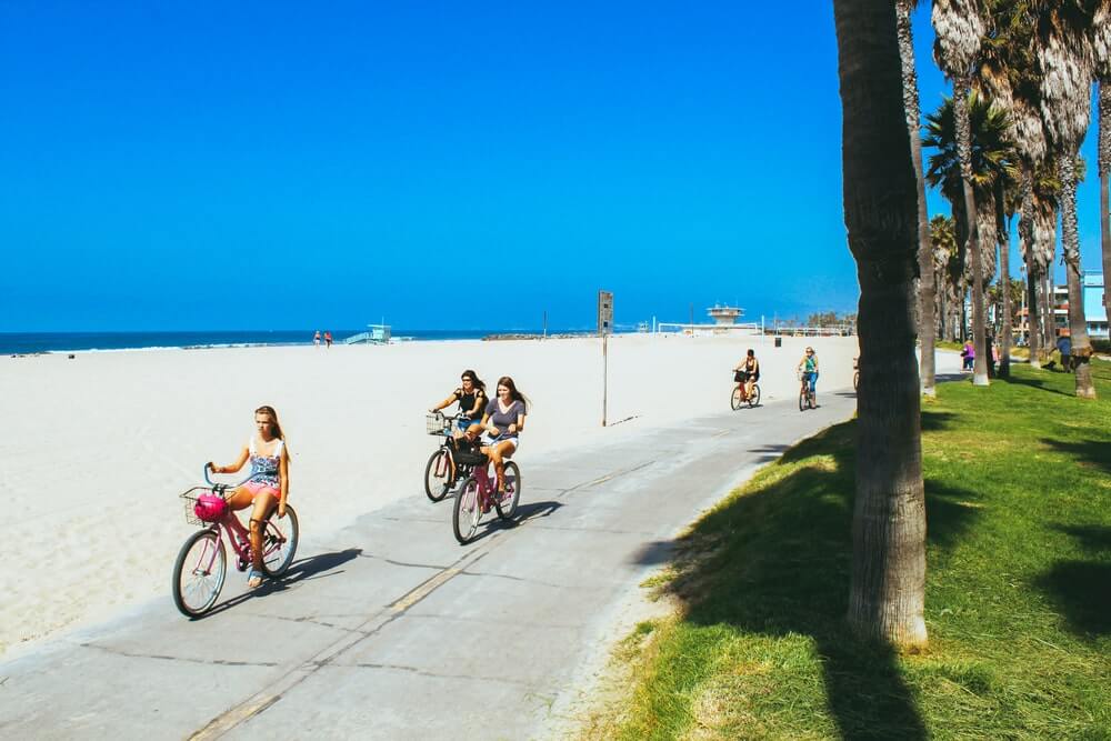 A group of people wearing light clothing biking along a sandy expanse by the water.